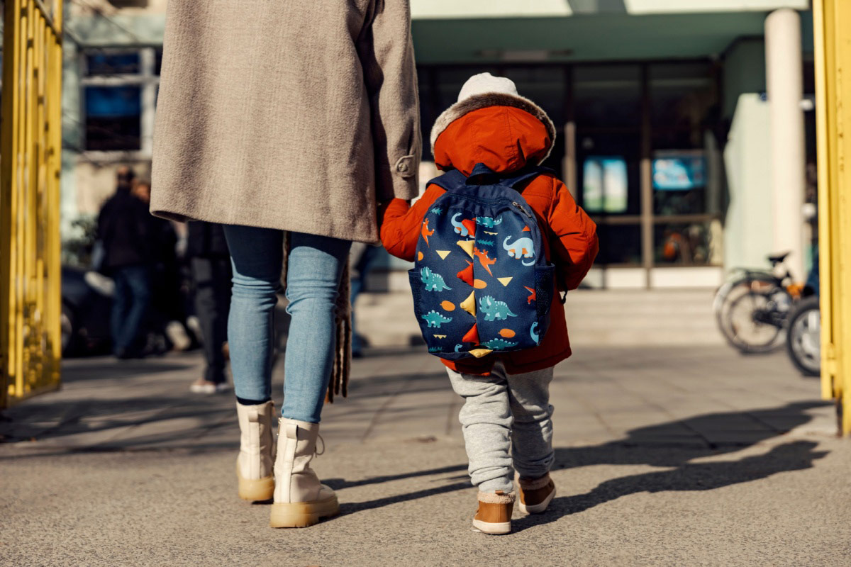 A parent and child walking into a school building, the child's backpack prominently featured.