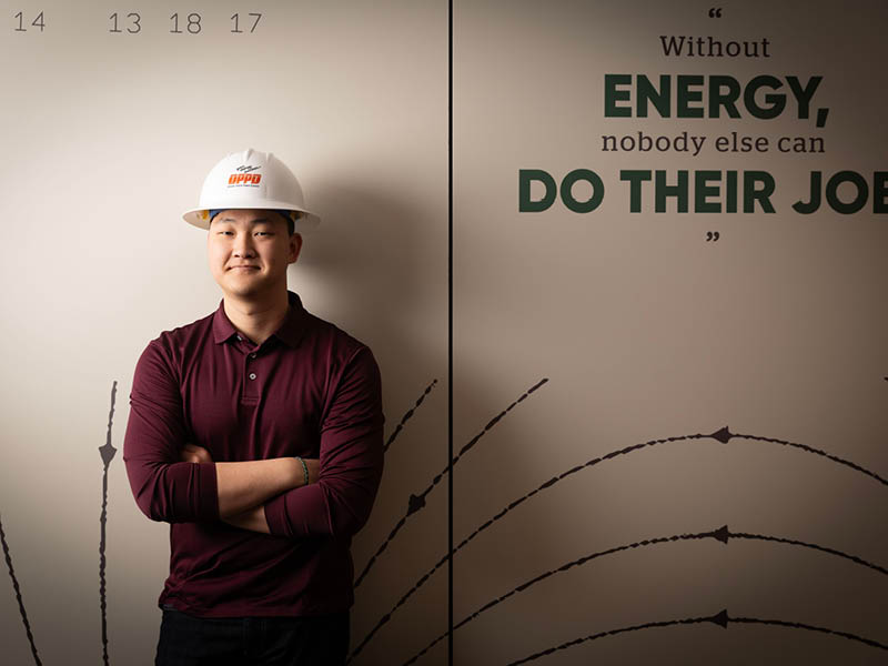 Student wearing a hard hat stands in front of a wall covered in engineering diagrams.
