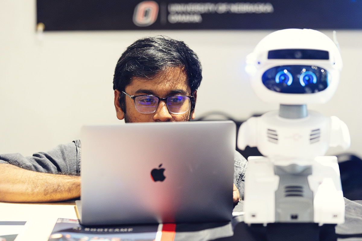 A student works on a laptop while a small robot sits next to the computer.