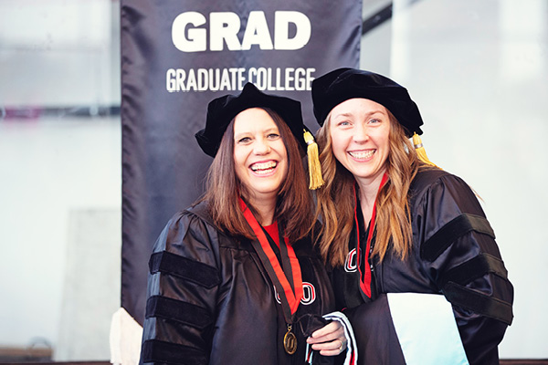 two graduate students are smiling for a photo in their regalia at graduation