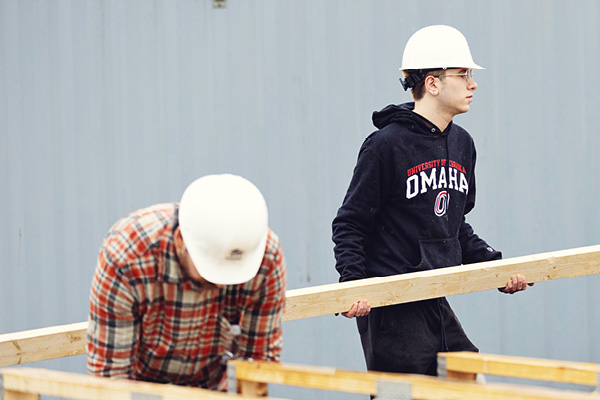 A man carrying a two by four wearing a hard hat.