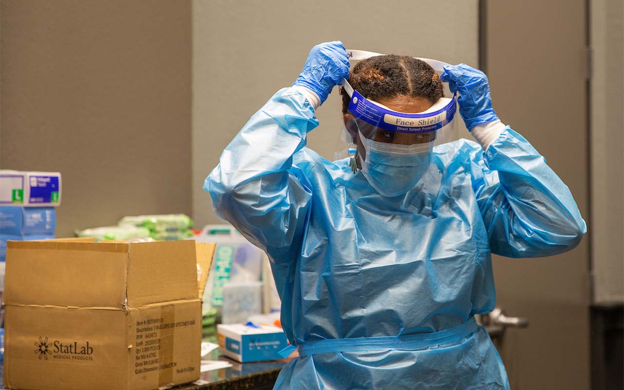 A woman in protective gear puts on a face shield as she prepares to collect samples for COVID-19 testing at UNO's on-campus testing site.