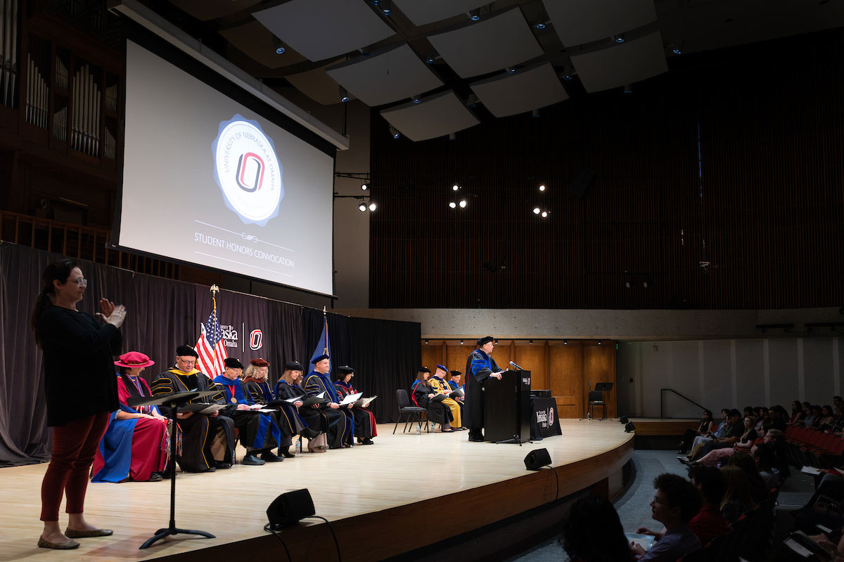 A speaker addresses the audience at the 2025 Student Honors Convocation held at the University of Nebraska at Omaha's Strauss Concert Hall. Faculty members, dressed in academic regalia, are seated on stage, and a sign language interpreter is visible on the side.
