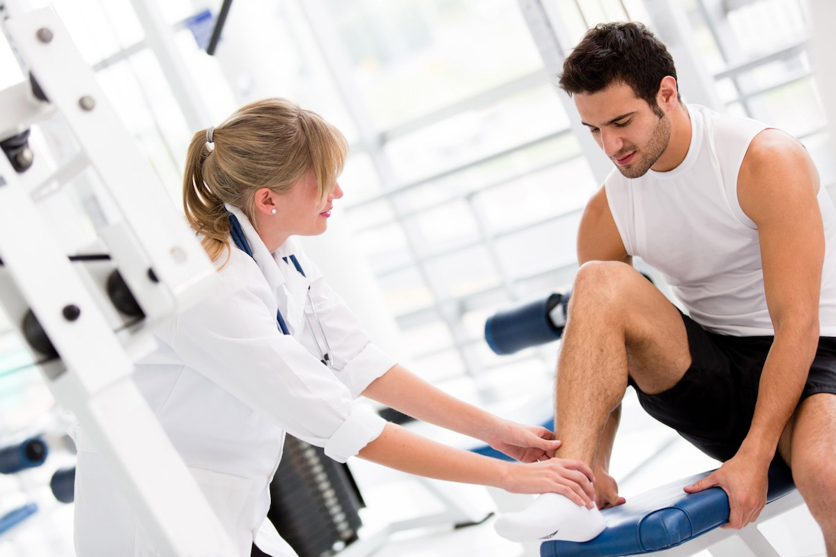 The image shows a healthcare professional, a woman wearing a white lab coat and stethoscope, assisting a man in athletic wear with his leg during a physical therapy session in a gym-like environment.