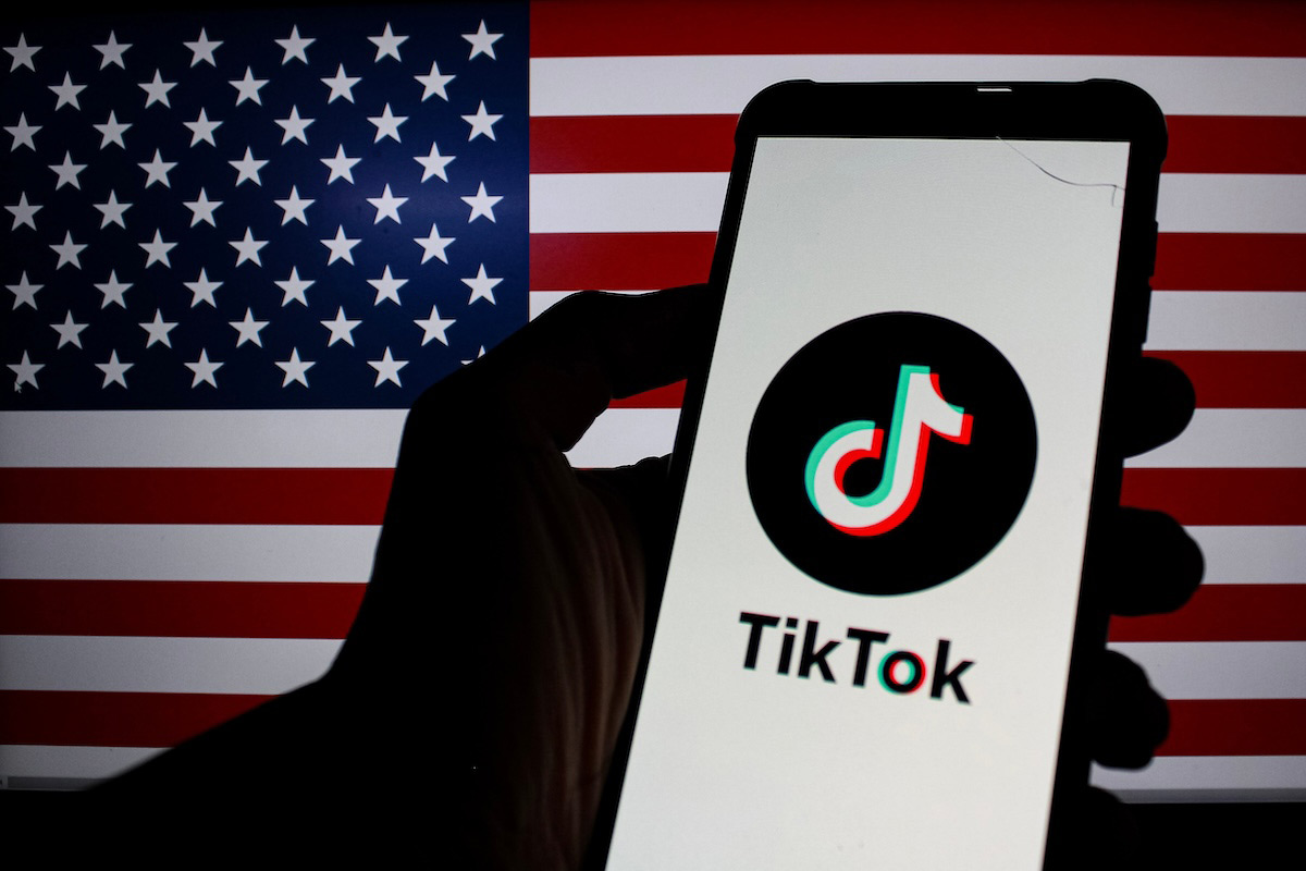 A phone showing the TikTok app is held in front of an American flag.