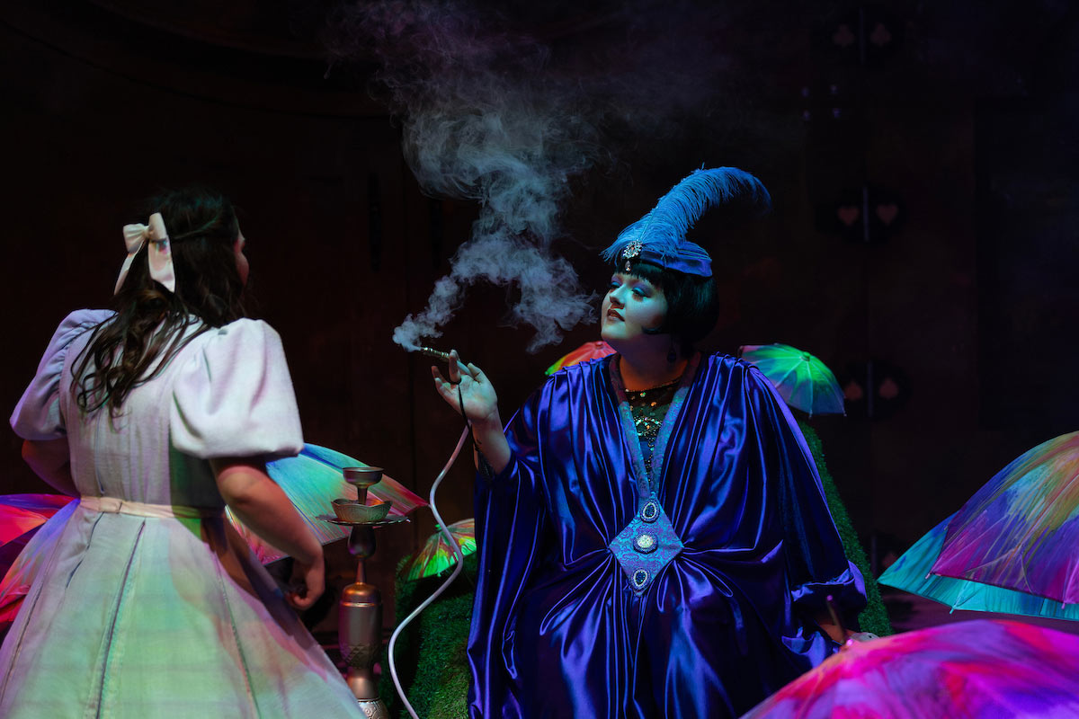 An actor in a vibrant blue costume with a feathered headpiece plays the Caterpillar, blowing smoke from a hookah while speaking to Alice on a colorful, glowing set.