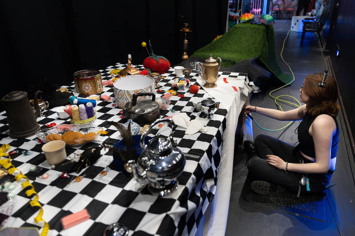 A crew member crouches beside a long table covered in colorful props—teapots, spools of thread, cups, and candy—prepping a tea party scene backstage for Alice in Wonderland.