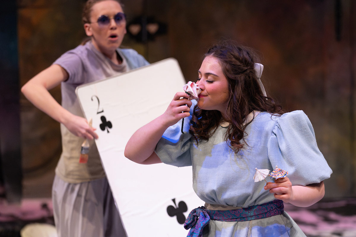 An actor playing Alice smells a whimsical paper flower on stage, holding tiny cocktail umbrellas, while another actor dressed as the 2 of Clubs looks on in surprise.
