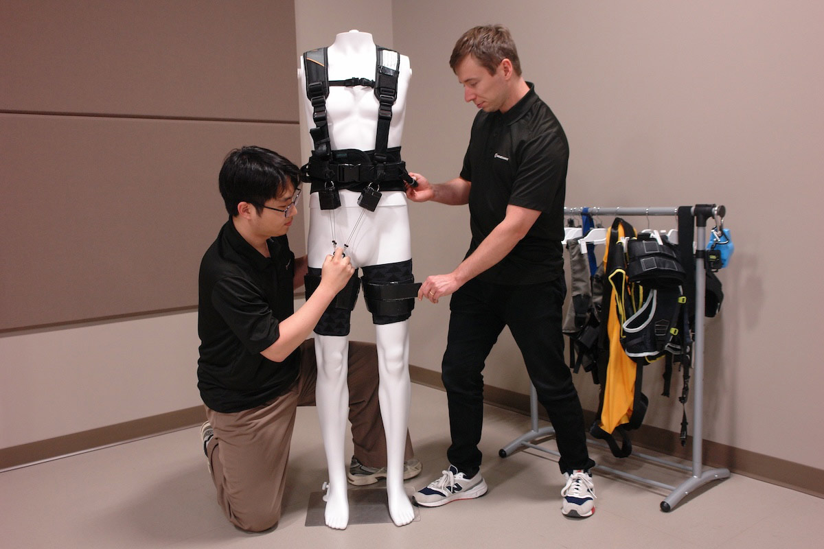 Two researchers work with an exoskeleton suit on a mannequin. One is adjusting the leg straps, while the other secures the device, as they test the suit's potential for stroke rehabilitation.