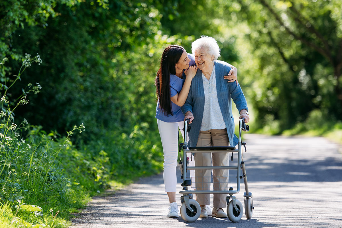Female caregiver and senior woman with walker on walk in nature. Nurse and elderly woman enjoying a warm day in nursing home, public park.