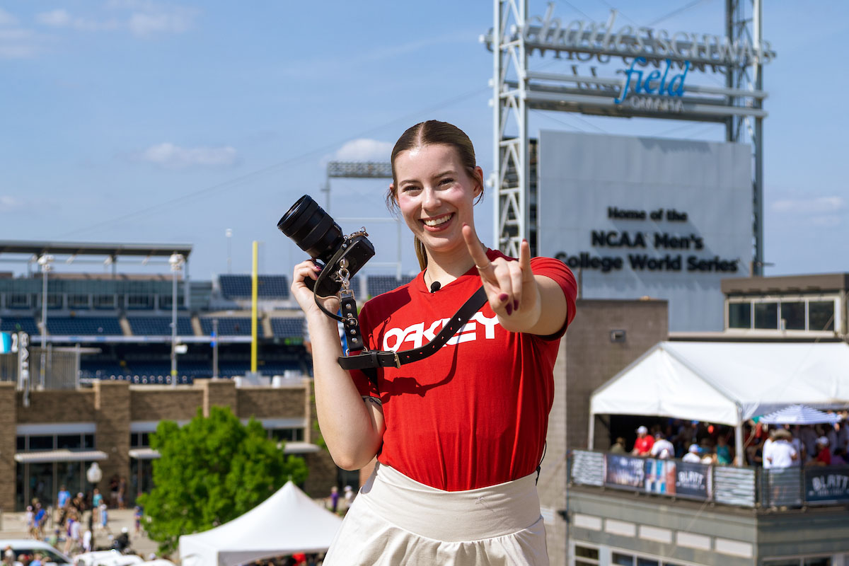 Bella McAtee smiles and makes the Mavericks horn symbol while holding a camera in front of Charles Schwab Field during the College World Series.