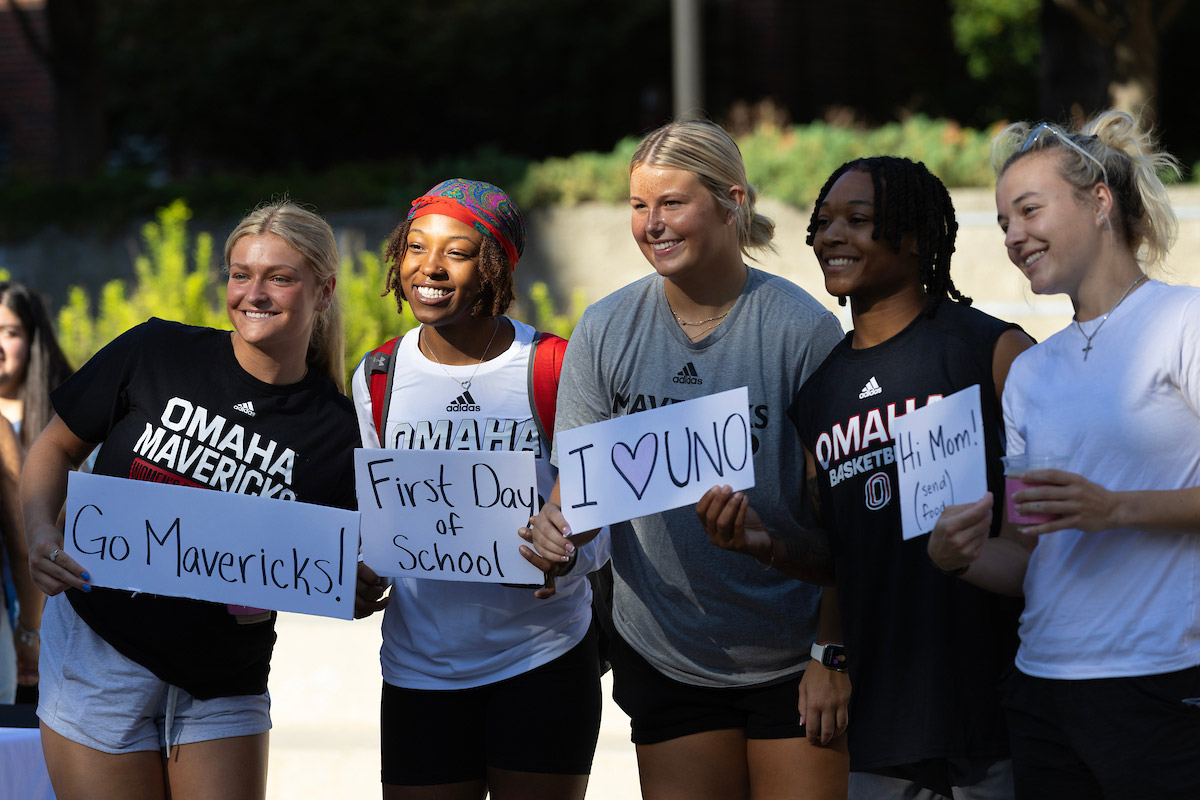 Five female university of Nebraska at Omaha students gather for a group photo on the first day of Fall 2024 classes in the Milo Bail Student Center Plaza. Students are smiling and holding signs saying “Go Mavericks,” First Day of School, “I heart UNO,” and “Hi Mom (send food)”