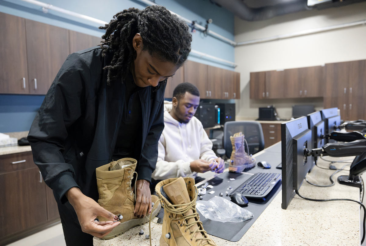 Two students work in a lab setting outfitting tan military-style boots with small electronic sensors. One student focuses on attaching a sensor to the sole of a boot, while the other assembles components at a nearby computer workstation.