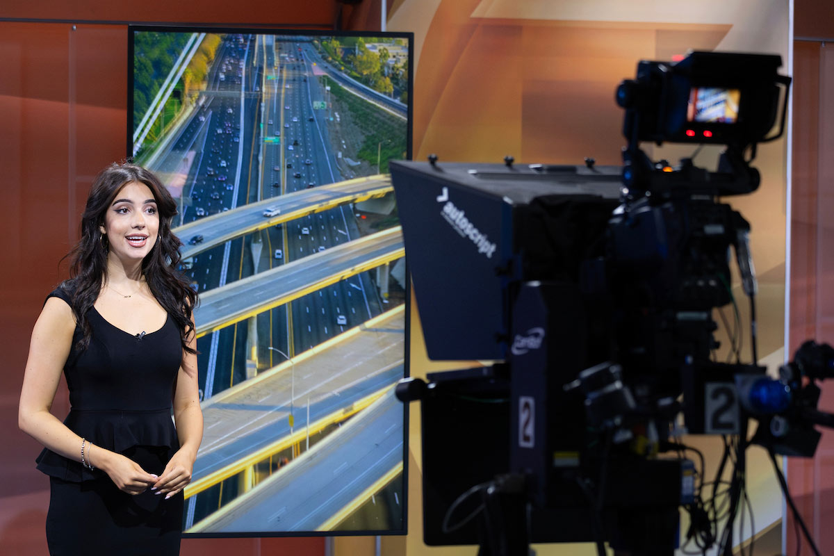Samantha Pastorino stands in a KETV television studio, speaking in front of a camera with an image of a highway interchange displayed on a screen behind her.