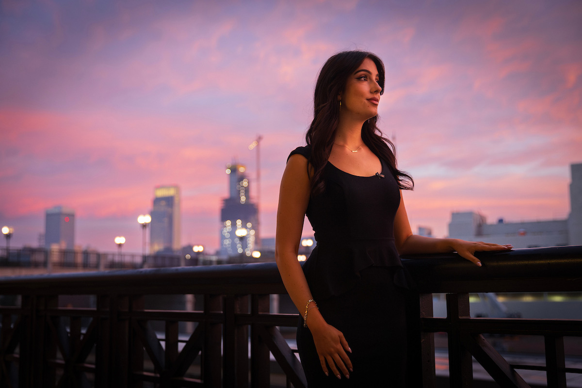 Samantha Pastorino, wearing a black dress, stands by a railing overlooking downtown Omaha at sunrise, with a pink and purple sky behind her.