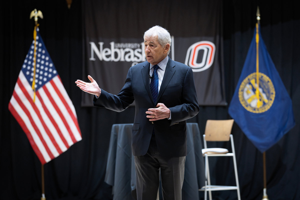 Chuck Hagel stands on stage speaking to an audience at the University of Nebraska at Omaha during the Chuck Hagel Symposium in Public Service, with the American and Nebraska state flags behind him.