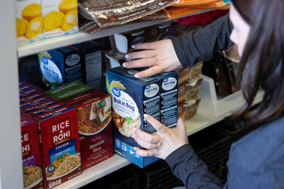A person organizes food items on a pantry shelf, placing boxes of Great Value Boil-in-Bag white rice next to boxes of Rice-A-Roni and Hy-Vee Spanish Rice. The shelves are stocked with various boxed and packaged food products. 