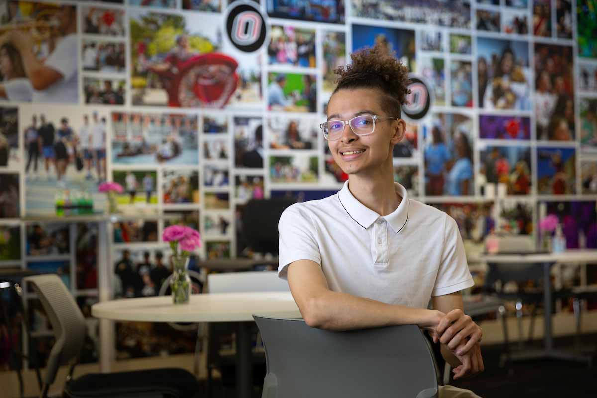 UNO student Dylan Clute smiles while wearing glasses and a white polo shirt sits in a modern room with tables, chairs, and vases of pink flowers. The background features a collage wall filled with colorful photos of campus life and the University of Nebraska at Omaha “O” logo. 