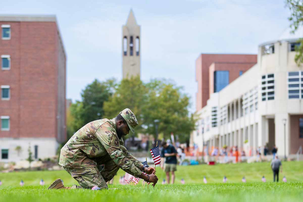 A person in military uniform kneels on a grassy area to place small American flags in the ground. 