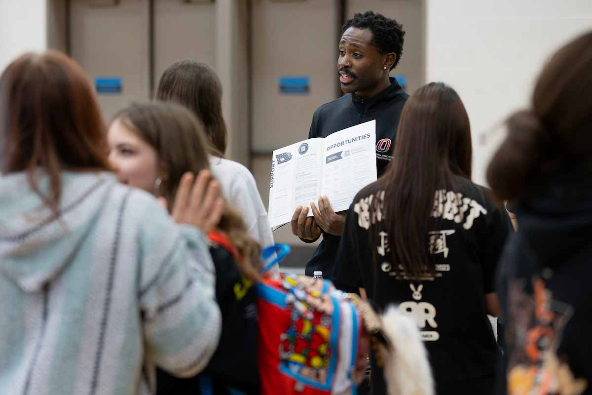 Fabio Gbetanou, a recruiter for the University of Nebraska at Omaha, speaks with a prospective student at a high school career fair. He holds an informational packet while standing behind a UNO display table covered with brochures and a large UNO logo.