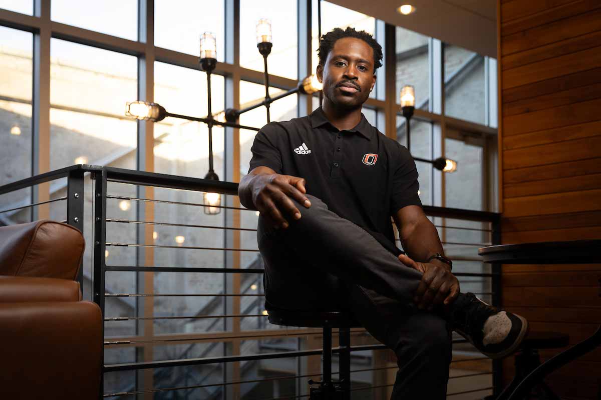 Fabio Gbetanou, a recruiter for the University of Nebraska at Omaha, sits confidently in a modern, well-lit indoor space with large windows and industrial-style lighting, wearing a black UNO polo shirt and looking toward the camera. 