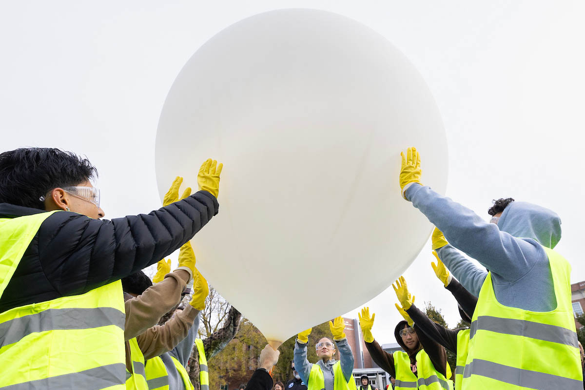 A group of people wearing yellow safety vests and gloves gathered outdoors, working together to hold a large, white balloon. The scene captures their collaboration in a science or educational project under overcast skies.