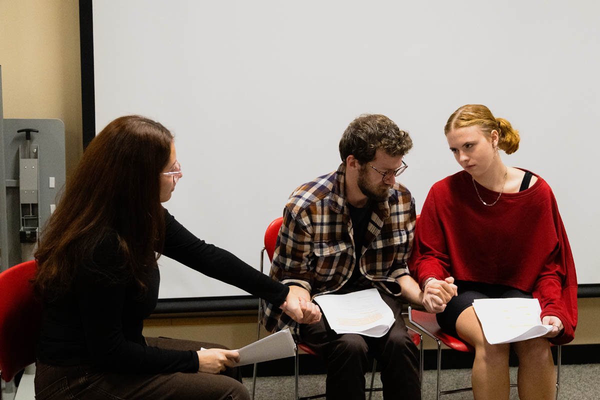 Three people sitting in a row, holding hands and reading scripts in a rehearsal setting, with serious expressions.