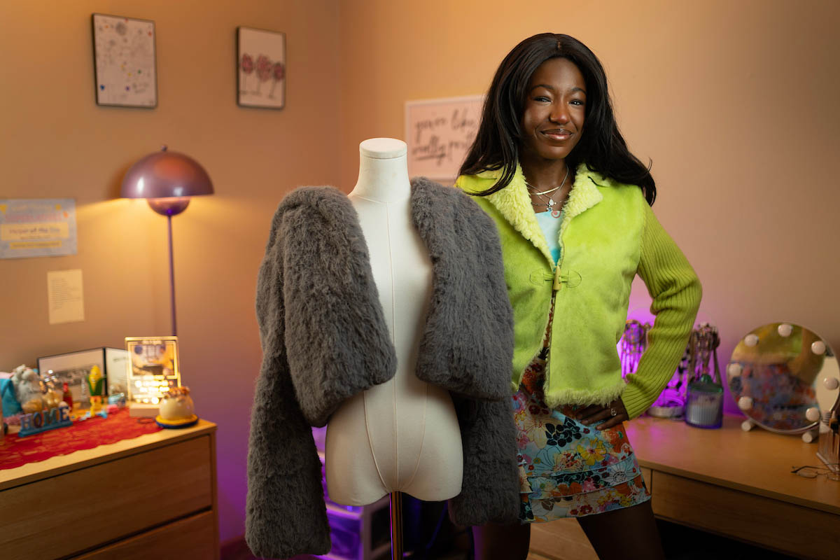 Lydia stands beside a mannequin displaying a gray fur jacket. She wears a bright green jacket and a colorful floral skirt, posing in a well-lit, cozy room with various decorative items on the dresser.