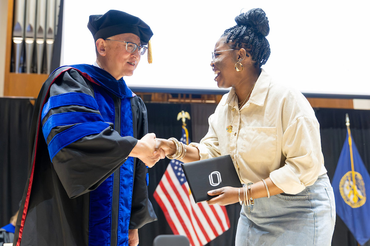 A university official in academic regalia shakes hands with a smiling student holding a UNO-branded award folder on stage during a formal honors ceremony, with U.S. and Nebraska flags in the background. 