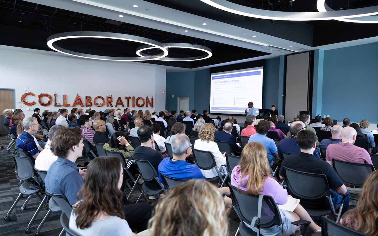 A large audience sits in a CEC conference room facing a presentation screen while a speaker addresses the crowd from the front. The word “COLLABORATION” is displayed in large letters on the wall. 