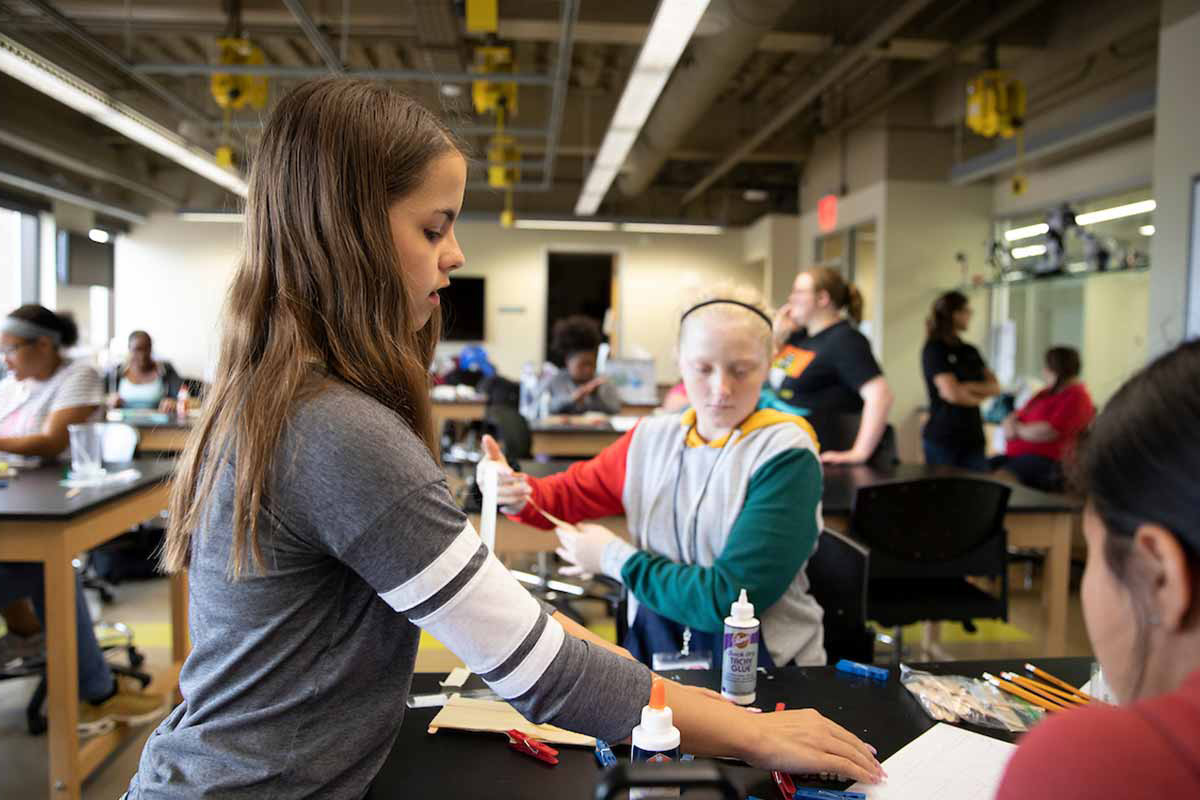 A young female student stands at a desk in a science lab, reaching for materials while speaking to her fellow classmates. 