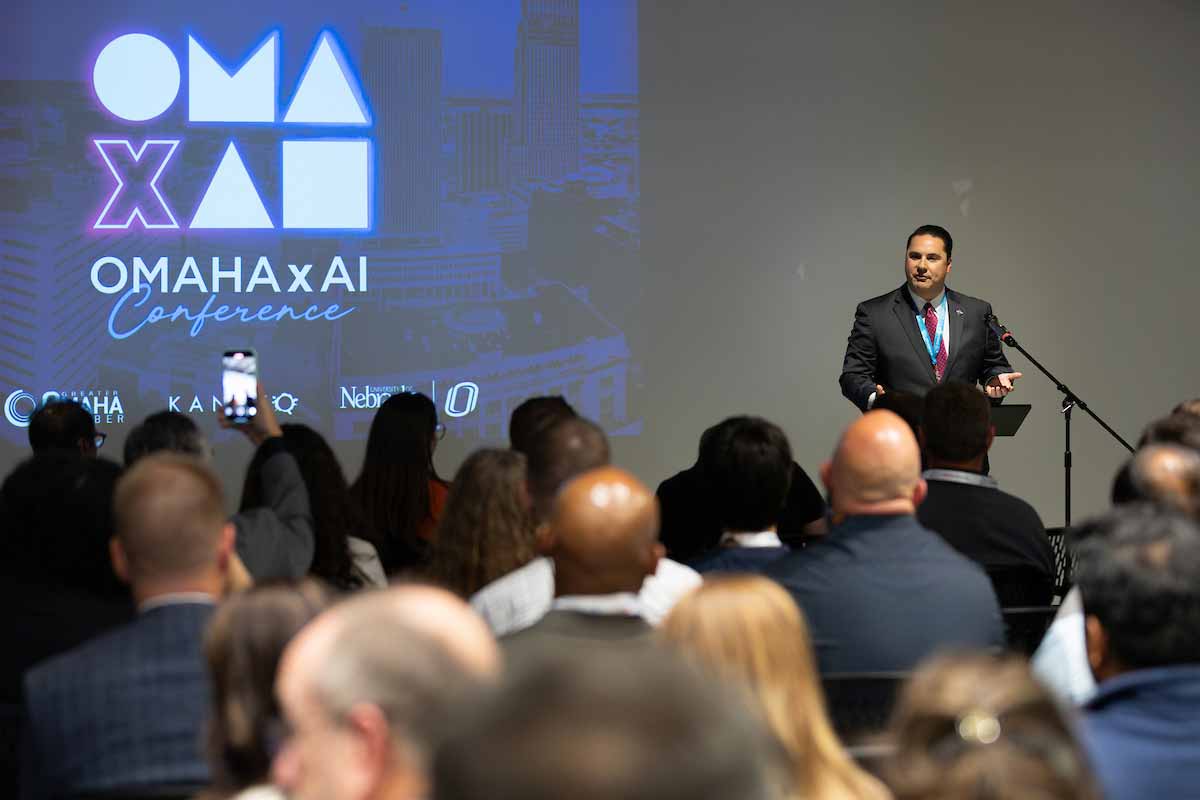 An audience listens as a speaker welcomes attendees during the OMA x AI Conference opening session, with the conference logo projected on a large screen behind the stage. 