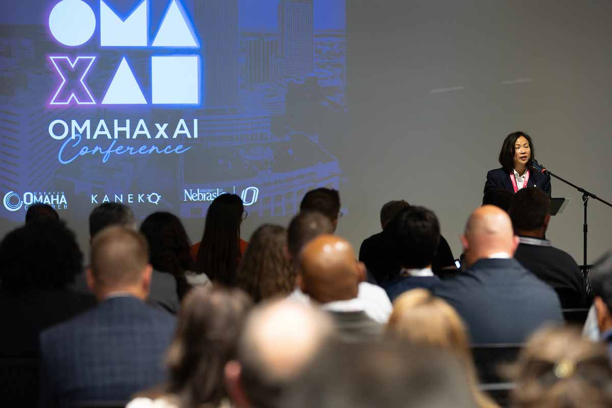 An audience listens as a speaker welcomes attendees during the OMA x AI Conference opening session, with the conference logo projected on a large screen behind the stage. 
