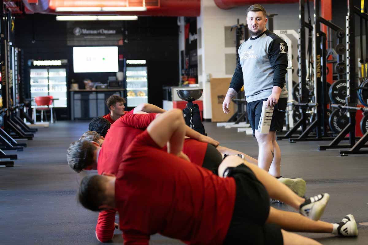 A student worker watches on as a row of athletes perform stretches at a weight room.  