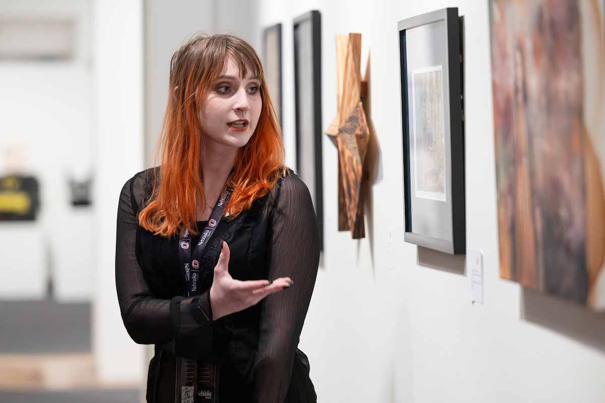 Student gestures toward framed artwork displayed on a gallery wall at UNO’s Student Research and Creative Activity Fair. 
