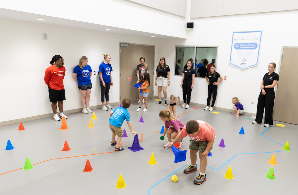 Young children navigate a colorful obstacle course with cones and markers while UNO student interns stand around the room encouraging and supervising the activity.