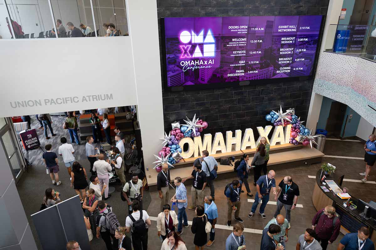 Attendees gather and network in the Union Pacific Atrium during the Omaha x AI Conference, beneath a large screen displaying the event schedule and an illuminated “OMAHA X AI” sign.