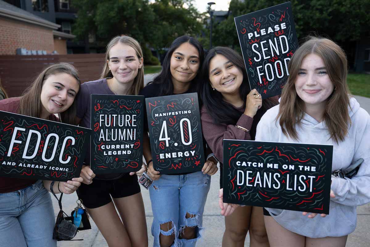 Five UNO students smile and hold signs with messages including “Future Alumni,” “Manifesting 4.0 Energy,” and “Catch Me on the Dean’s List” during an outdoor campus event. 