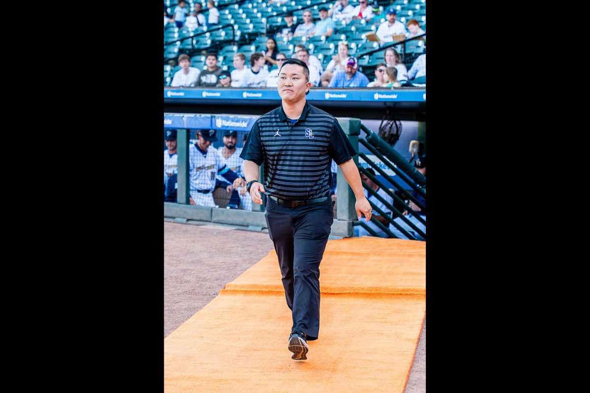 A man a black striped polo shirt and dark pants, walks down an orange carpet on the field at a baseball stadium. Players stand in the dugout behind them, and spectators fill the stadium seats in the background.