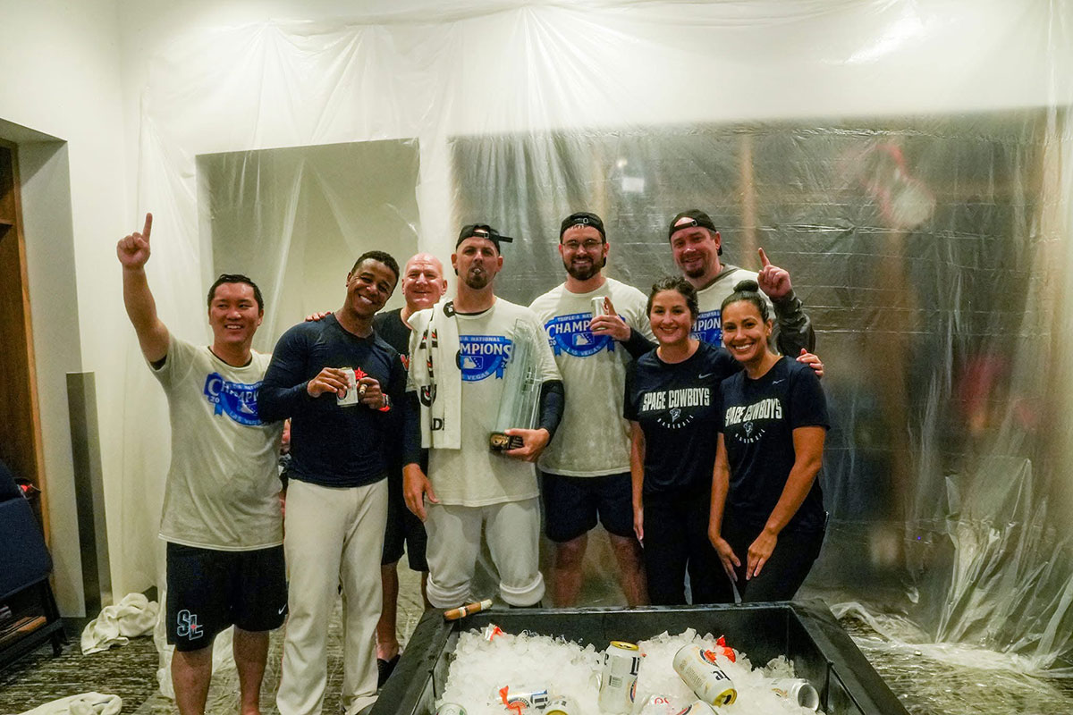 A group of nine people pose together during a postgame locker room celebration, with several wearing white championship T-shirts. Clear plastic sheeting covers the walls behind them, and a large tub filled with ice and drinks sits in the foreground. 