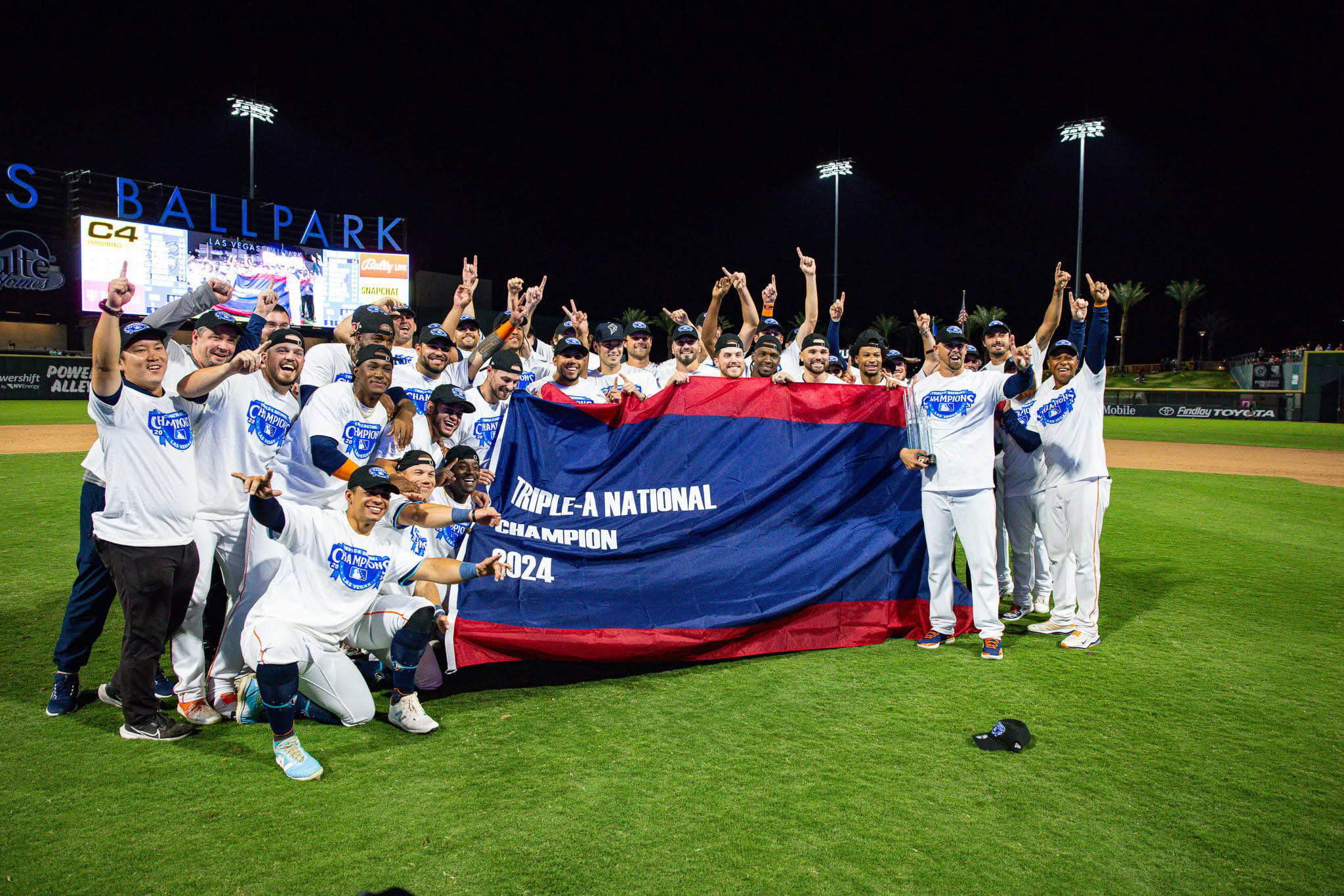 A baseball team celebrates on the field at night, posing together in white commemorative shirts and holding a large banner that reads “Triple-A National Champion 2024.” Players smile, point upward, and raise their hands in celebration while standing on the grass in front of the scoreboard at Las Vegas Ballpark 