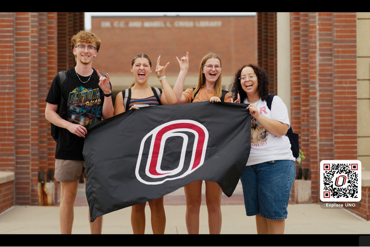 Four University of Nebraska at Omaha students stand outdoors in front of the Dr. C.C. and Mabel L. Criss Library, smiling and holding a black UNO flag with the Maverick “O” logo.