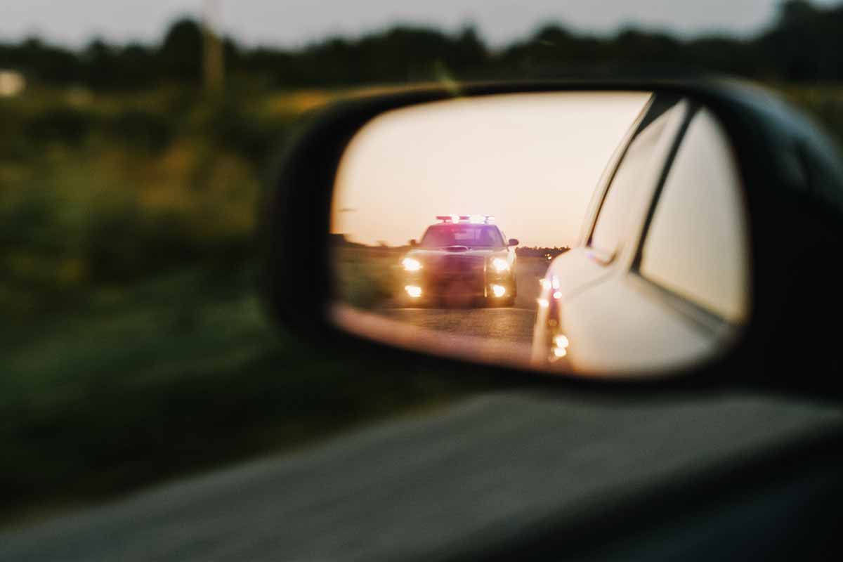 A car’s side mirror reflects a police vehicle with flashing red and blue lights following closely behind on a rural road at sunset 