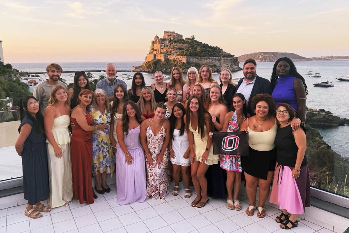 A group holds a UNO flag while standing in front of an Italian island.