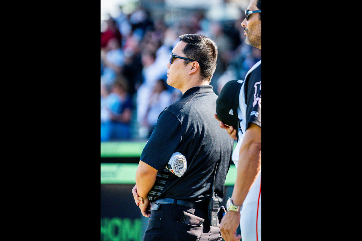 A man in a black striped polo shirt and dark pants faces away from the camera with his hands crossed in front of him and a rolled towel under his arm. A baseball player with his hand and hat over his chest stands in the foreground.  