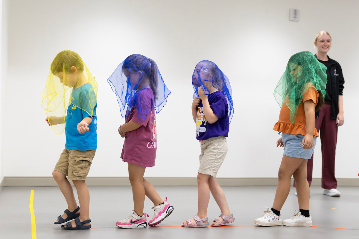Four young children wearing yellow, blue, and green mesh scarves over their heads walk in a line across a room during a movement or sensory activity. A UNO student intern stands in the background, smiling as they observe.