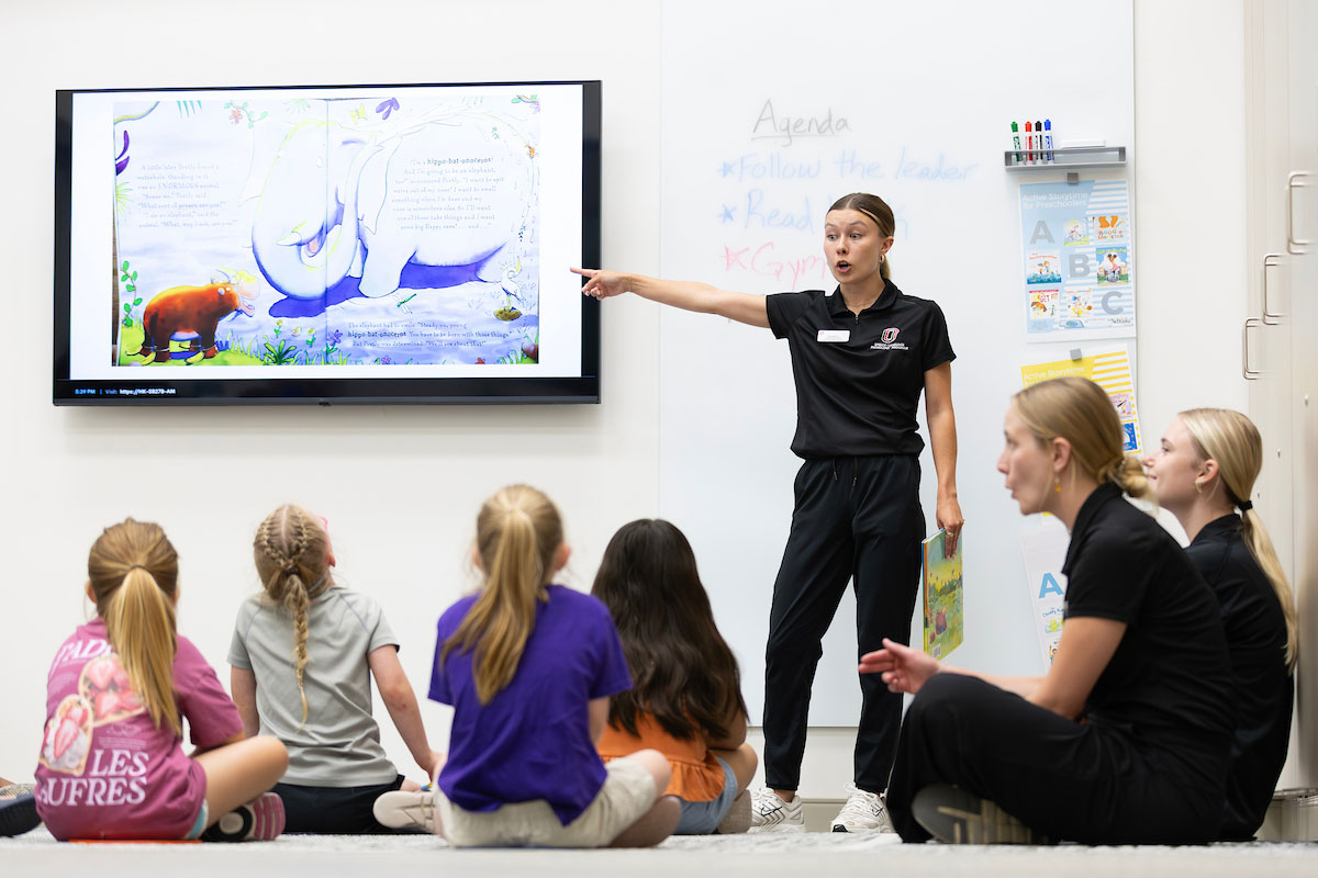 A UNO student intern reads aloud to a group of young children seated on the floor while pointing to illustrations displayed on a large screen.