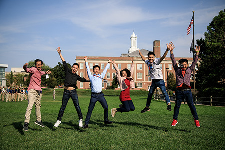 Five students are jumping and smiling on a green lawn in front of a campus building with a white cupola. The sky is clear and blue, and an American flag waves in the background. The scene captures a joyful, energetic moment on campus.