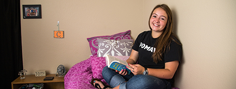 A UNO student sitting in her on-campus housing apartment, reading a book, and smiling at the camera.