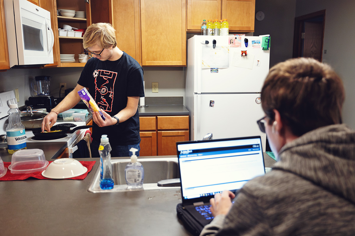A student cooks in a residence hall kitchen while another student studies on a laptop.